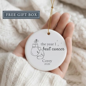 May include: White ceramic ornament with a gold string, featuring a boxing glove design and the text "the year I beat cancer" with a name and the year 2026. The ornament is held in a hand.