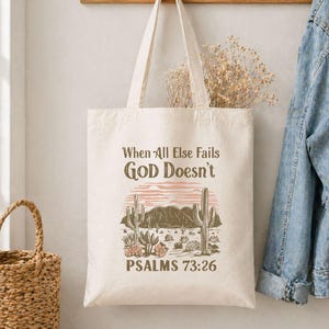 May include: A cream-colored canvas tote bag featuring a desert landscape graphic with cacti and mountains. The text reads When All Else Fails GOD Doesn't and PSALMS 73:26 in brown lettering. The design includes a soft pink sunset background.