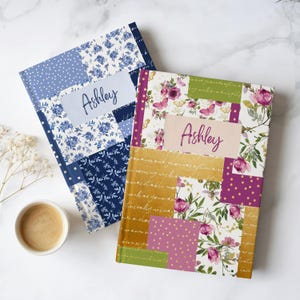 May include: Two personalized journals with the name "Ashley" on the cover. One journal features a patchwork design with blue floral patterns, while the other has a floral and butterfly design with pink and gold accents. A cup of coffee sits nearby.