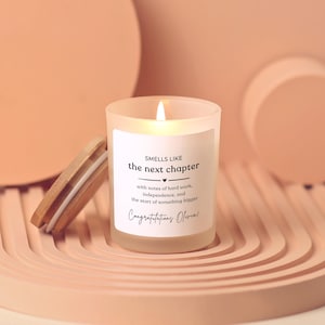 May include: A frosted glass candle with a lit wick and a bamboo lid. The white label features the text: SMELLS LIKE the next chapter, with notes of hard work, independence, and the start of something bigger. Congratulations Olivia!