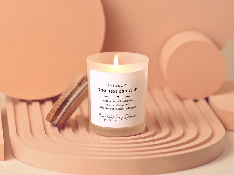 May include: A frosted glass candle with a lit wick and a bamboo lid. The white label features the text: SMELLS LIKE the next chapter, with notes of hard work, independence, and the start of something bigger. Congratulations Olivia!