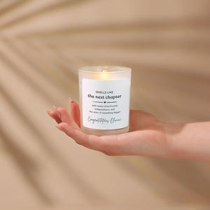 May include: A frosted glass candle with a white label featuring the black text: SMELLS LIKE the next chapter, with notes of hard work, independence, and the start of something bigger, Congratulations Olivia. The candle has a visible warm glow.