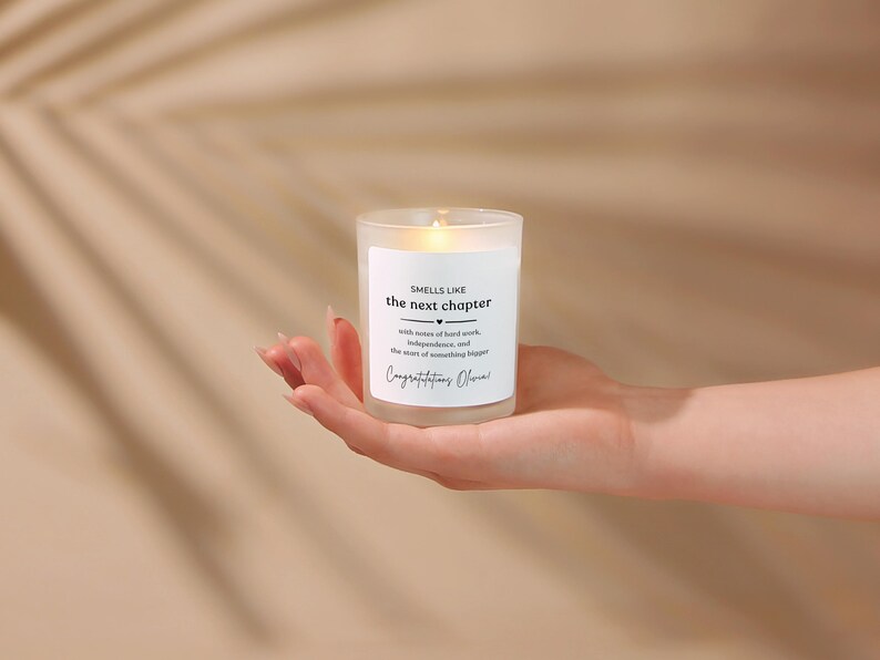 May include: A frosted glass candle with a white label featuring the black text: SMELLS LIKE the next chapter, with notes of hard work, independence, and the start of something bigger, Congratulations Olivia. The candle has a visible warm glow.