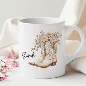 May include: A white ceramic mug featuring a printed illustration of two light brown cowboy boots adorned with a bouquet of pink and cream-colored roses and greenery. The name Sarah is printed in black cursive script to the left of the boots.
