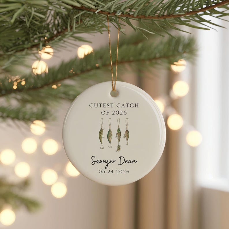 May include: A round white ceramic ornament hanging by a gold string. The text reads CUTEST CATCH OF 2026, Sawyer Dean, and 05.24.2026. Below the first line of text are four small, detailed illustrations of fishing lures in green and brown tones.