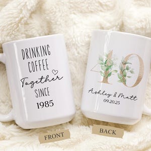 May include: Two white ceramic mugs. One mug says "DRINKING COFFEE Together SINCE 1985". The other mug has the number "40" with floral accents and the names "Ashley & Matt" and the date "09.20.25".