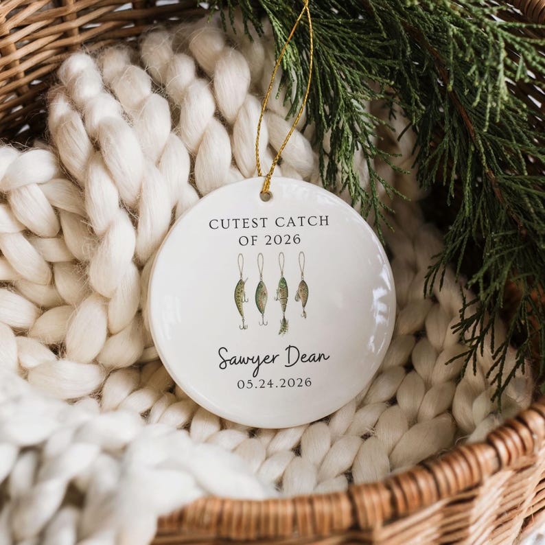 May include: A round white ceramic ornament featuring a gold hanging string. The text reads CUTEST CATCH OF 2026, Sawyer Dean, and 05.24.2026. Below the top text are four illustrated green fishing lures with hooks hanging from thin lines.
