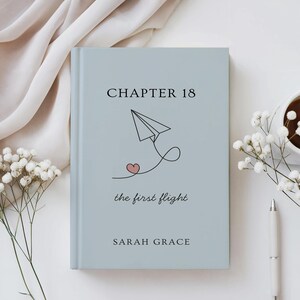 May include: A light blue hardcover book with the title "Chapter 18" and the phrase "the first flight." A paper airplane illustration with a heart is featured on the cover, along with the name "Sarah Grace." The book is surrounded by white flowers, a pen, and a cup of coffee.