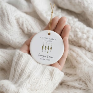 May include: A round white ceramic ornament featuring the text CUTEST CATCH OF 2026, an illustration of four fishing lures, and the name Sawyer Dean with the date 05.24.2026 printed in black, suspended by a thin gold metallic hanging cord.