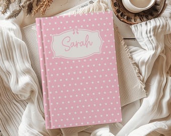 May include: A pink journal with white polka dots and the name "Sarah" in a decorative frame. The journal is hardcover and rests on a neutral-colored cloth, with a candle and decorative elements in the background.