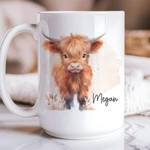 K&ouml;nnte beinhalten: Wei&szlig;e Keramiktasse mit einer Aquarellillustration einer Hochlandkuh. Die Kuh ist in Braun- und Oranget&ouml;nen gehalten, mit dem Namen "Megan" in schwarzer Schrift. Die Tasse hat einen gro&szlig;en Henkel.