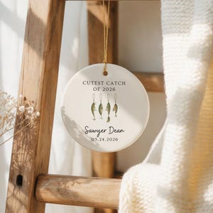 May include: A round white ceramic ornament hanging by a gold string. The text reads CUTEST CATCH OF 2026, Sawyer Dean, and 05.24.2026. Below the first line of text are four small, detailed illustrations of fishing lures in green and brown tones.