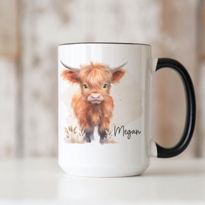 K&ouml;nnte beinhalten: Wei&szlig;e Keramik-Tasse mit schwarzem Griff und Rand. Die Tasse zeigt eine Aquarell-Illustration einer Hochlandkuh mit braunem Fell und H&ouml;rnern. Der Name "Megan" ist in Schreibschrift auf die Tasse geschrieben.