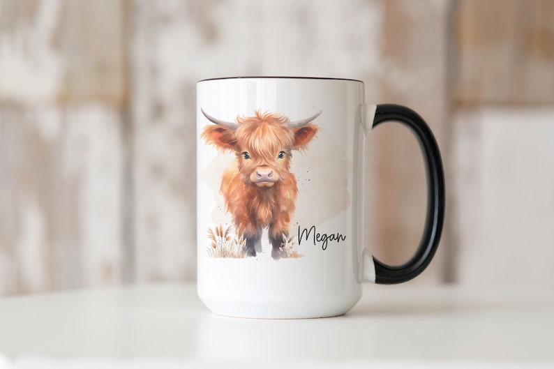 K&ouml;nnte beinhalten: Wei&szlig;e Keramik-Tasse mit schwarzem Griff und Rand. Die Tasse zeigt eine Aquarell-Illustration einer Hochlandkuh mit braunem Fell und H&ouml;rnern. Der Name "Megan" ist in Schreibschrift auf die Tasse geschrieben.