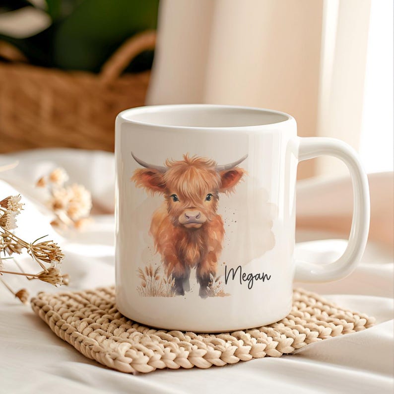 K&ouml;nnte beinhalten: Wei&szlig;e Keramiktasse mit einer Aquarellillustration einer Hochlandkuh mit langem, zotteligem, rotbraunem Fell und gebogenen H&ouml;rnern. Der Name "Megan" ist kursiv geschrieben. Die Tasse steht auf einem beigefarbenen, gewebten Untersetzer.