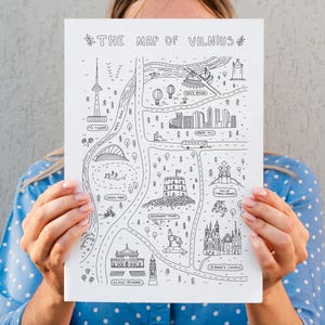 Puede incluir: Un mapa ilustrado en blanco y negro de Vilna, Lituania, sostenido frente a una persona. El mapa presenta puntos de referencia, carreteras y el texto "THE MAP OF VILNIUS". El mapa está detallado con árboles, edificios y otros elementos.
