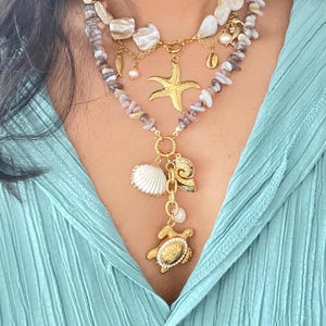 Peut inclure: Un collier superposé avec un pendentif étoile de mer doré, des breloques coquillages et un pendentif tortue. Le collier comprend des perles de pierres naturelles, des perles et des accents de coquillages. Le collier est porté sur une chemise bleu clair.