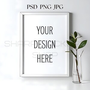 May include: A white framed art print mockup with the text "YOUR DESIGN HERE". A clear glass vase with a green leafy stem is to the right of the frame. The text "PSD PNG JPG" is at the top.