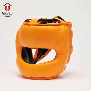 Op de afbeelding: Oranje leren bokshoofdbescherming met zwarte binnenvoering. De hoofdbescherming heeft een grote opening voor de ogen en neus, en een band aan de bovenkant. Het logo "UNDER SPORTS CLUB HOUSE" staat in de linkerbovenhoek.