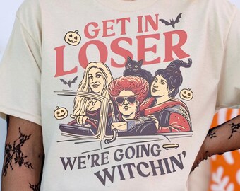 May include: Beige t-shirt with a Halloween-themed graphic. The design features the text "GET IN LOSER WE'RE GOING WITCHIN'" above an illustration of three witches in a car. Bats and pumpkins are also included in the design.