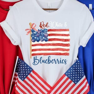 May include: Three t-shirts in red, white, and blue. The white t-shirt has a watercolour design of an American flag made of blueberries. The flag reads "Red, White & Blueberries". Two small American flags are in front.