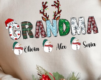 May include: A cream-colored sweatshirt with the word "GRANDMA" in a festive, patterned design. The letter "G" has a Santa hat, and the letter "A" has reindeer antlers. Below the word are three snowmen with the names Olivia, Alex, and Sofia.