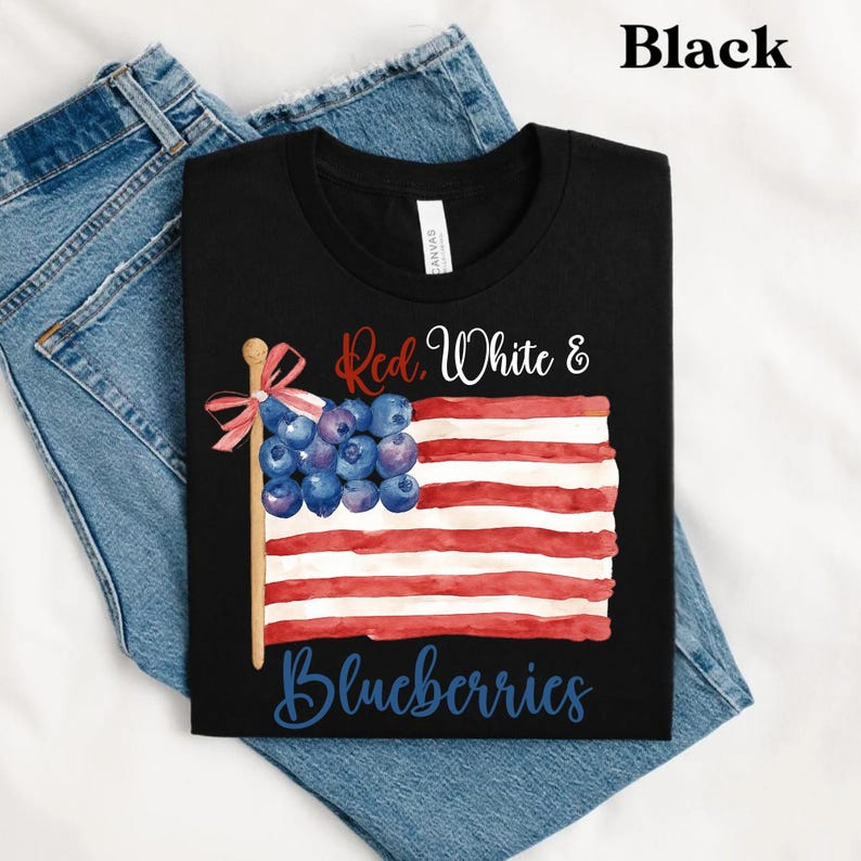 May include: Black t-shirt featuring a flag design made of red and white stripes and blueberries. The text on the shirt reads "Red, White & Blueberries". The t-shirt is folded on top of a pair of blue jeans.