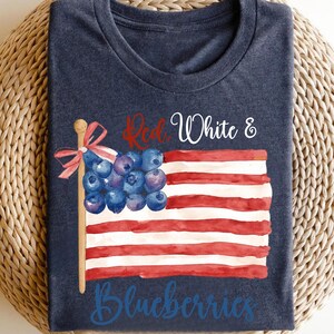 May include: A navy blue t-shirt with a watercolour design of the American flag. The flag's stars are replaced with blueberries, and the text "Red, White & Blueberries" is written across the design. The shirt is folded on a woven surface.