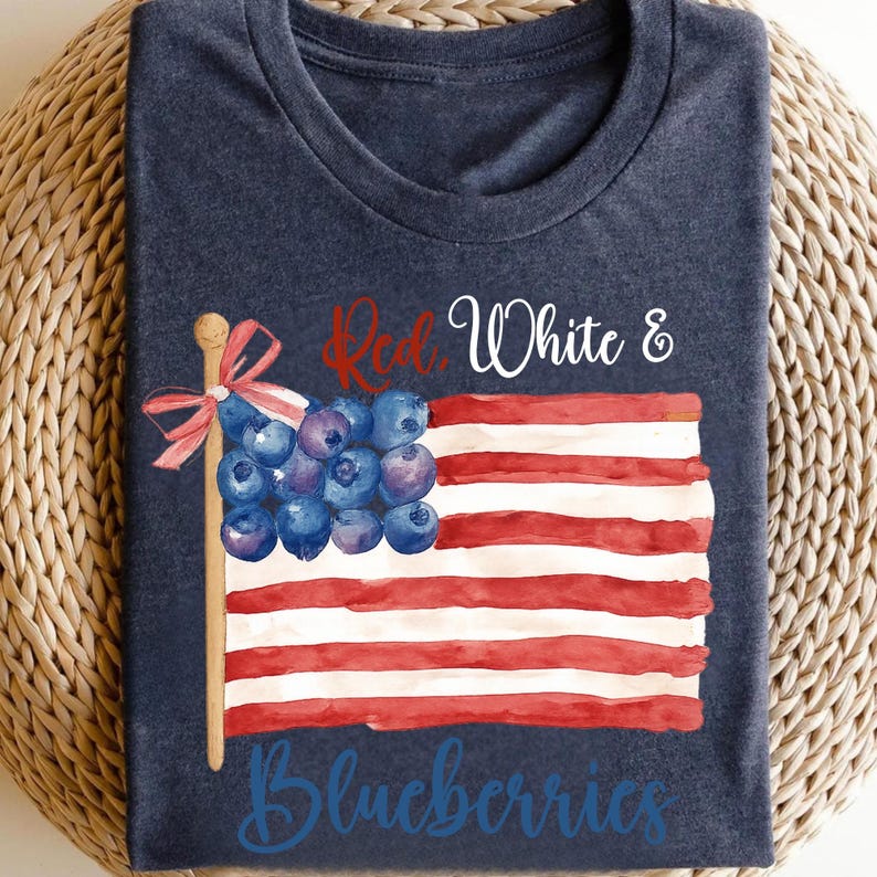 May include: A navy blue t-shirt with a watercolour design of the American flag. The flag's stars are replaced with blueberries, and the text "Red, White & Blueberries" is written across the design. The shirt is folded on a woven surface.