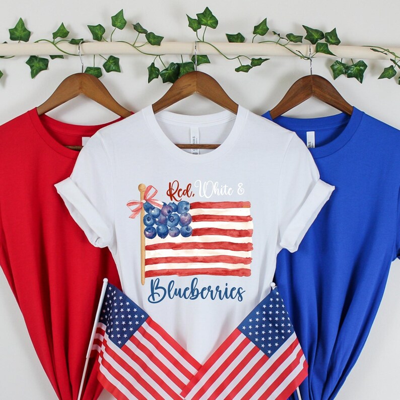 May include: Three t-shirts in red, white, and blue. The white t-shirt has a watercolour design of an American flag made of blueberries. The flag reads "Red, White & Blueberries". Two small American flags are in front.