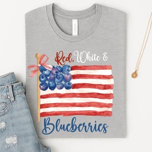 May include: A grey t-shirt with a watercolour design of an American flag made of red and white stripes and blueberries. The text "Red, White & Blueberries" is written across the design. A pair of blue jeans is partially visible.