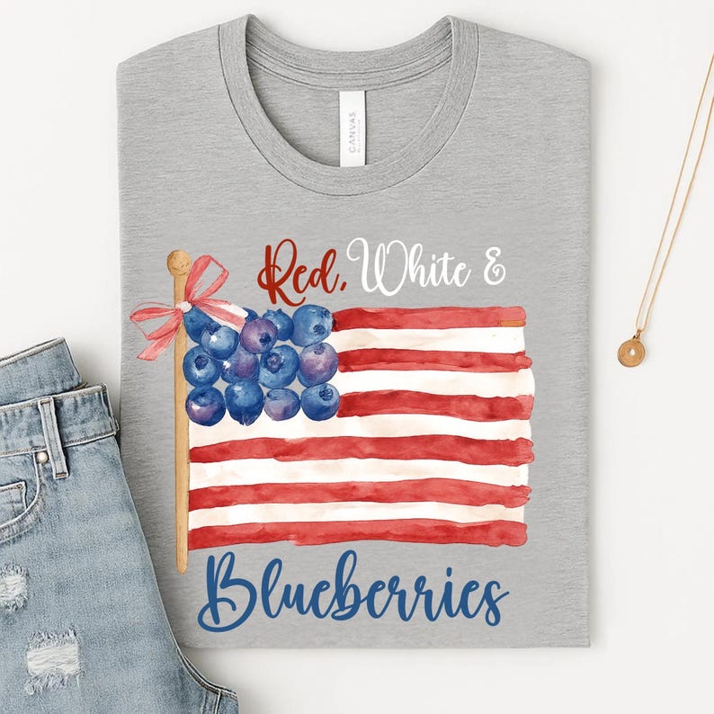 May include: A grey t-shirt with a watercolour design of an American flag made of red and white stripes and blueberries. The text "Red, White & Blueberries" is written across the design. A pair of blue jeans is partially visible.
