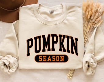 May include: Cream-colored sweatshirt with the words "PUMPKIN SEASON" in black and orange lettering. The sweatshirt has a crew neck and long sleeves. A brown hat and dried wheat stalks are in the background.