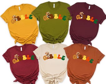 May include: Six short-sleeved t-shirts in various colors, including mustard yellow, olive green, and burgundy. Each shirt features the word "GOBBLE" in a colorful, retro font, with a turkey graphic. The shirts are likely for Thanksgiving.