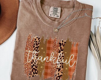 May include: A brown Comfort Colors t-shirt with a decorative design featuring leopard print, plaid, and glitter accents. The word "thankful" is written in a cursive font across the design. A silver necklace with a pendant is draped across the shirt.