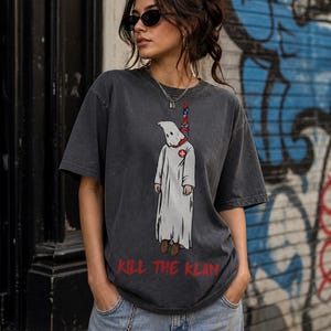 May include: Dark gray t-shirt with a graphic of a figure in a white robe and hood, with a red cross and a patterned hat. The text "KILL THE KLAN" is printed in red at the bottom. The person is wearing sunglasses and blue jeans.