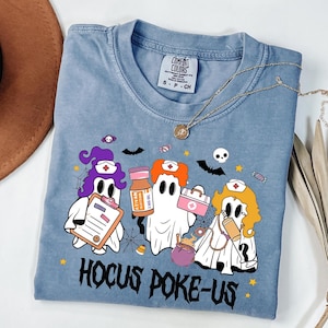 May include: A blue Comfort Colors t-shirt featuring a Halloween-themed graphic with ghost nurses, bats, and the text "Hocus Poke-Us." The ghosts hold medical supplies. A gold necklace is draped across the shirt.