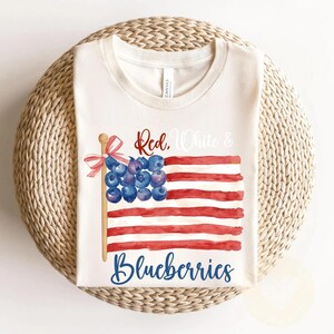 May include: A cream-coloured t-shirt with a watercolour design of an American flag made of red and white stripes and blueberries. The text "Red, White & Blueberries" is written across the design. The shirt is folded on a woven surface.