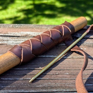 May include: A close-up of a wooden dowel wrapped with brown leather. A long, thin, gold-colored tool is lying next to the dowel. A brown leather strap is also visible.