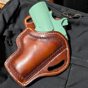 Könnte beinhalten: Ein braunes Lederholster mit einer hellgrünen Pistole darin. Das Holster ist so konzipiert, dass es eng an der Waffe anliegt und einen genähten Rand hat.