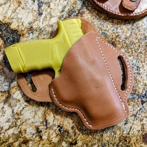May include: A brown leather holster with a yellow plastic handgun inside. The holster has white stitching and two belt loops.