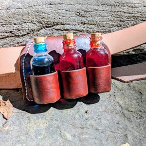 Puede incluir: Un cinturón de cuero marrón con un accesorio de funda que sostiene tres botellas de vidrio pequeñas. Las botellas están selladas con tapones de corcho y contienen líquidos en tonos vibrantes de azul, rojo brillante y un rojo más oscuro.