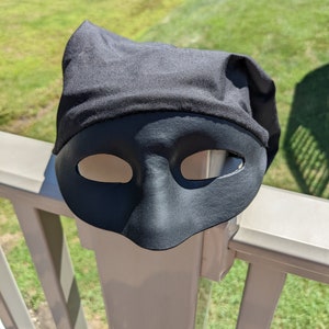 May include: A black masquerade mask with a fabric covering the top. The mask is sitting on a white railing.