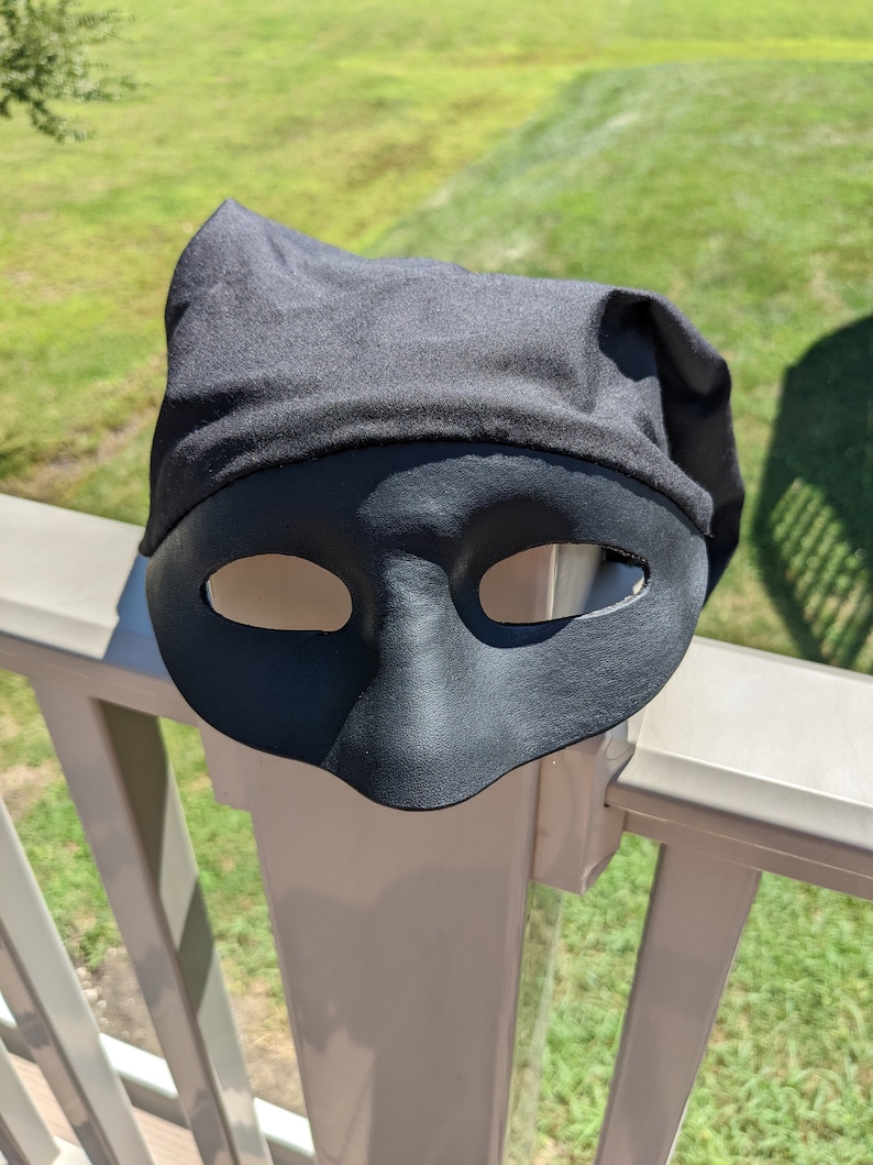 May include: A black masquerade mask with a fabric covering the top. The mask is sitting on a white railing.