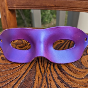 May include: A purple masquerade mask with two eye holes. The mask is made of a smooth, leather-like material and has a slight sheen.