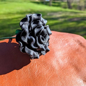 May include: A black leather rose with a green stem, resting on a brown leather surface.