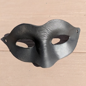 Pode incluir: Uma máscara de couro preto para um baile de máscaras com dois orifícios para os olhos. A máscara tem a forma de uma lágrima e tem uma parte superior ligeiramente curva.