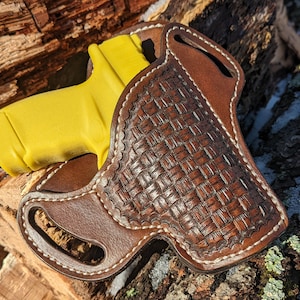 May include: A brown leather holster with a woven pattern and white stitching. The holster is designed to hold a handgun. The holster is sitting on a pile of wood.