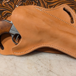 Puede incluir: Una funda de cuero marrón claro con costuras blancas, diseñada para un revólver. La funda cubre parcialmente el arma, que tiene una empuñadura de madera. El fondo presenta un diseño de cuero tallado.