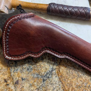 Pode incluir: Um coldre de couro marrom com costuras brancas para uma pistola. O coldre tem uma borda festonada decorativa.